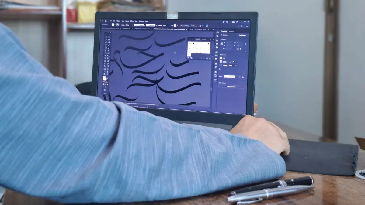 A point-of-view photo of a digital calligrapher at BinMahmood using a mouse and Adobe Illustrator to manually draw complex Arabic script strokes on a desktop computer.