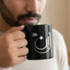 Black ceramic mug with “Zain” Arabic calligraphy held by bearded male model sipping coffee, face cropped below eyes, minimalist setting