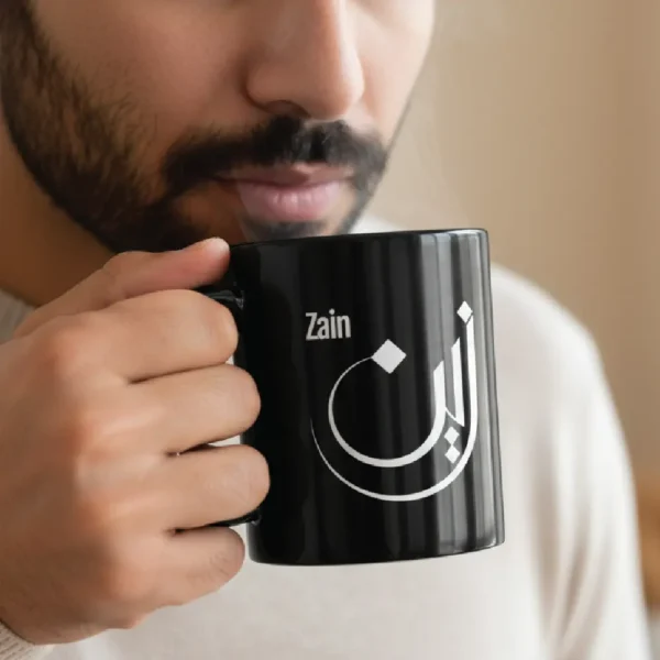 Black ceramic mug with “Zain” Arabic calligraphy held by bearded male model sipping coffee, face cropped below eyes, minimalist setting